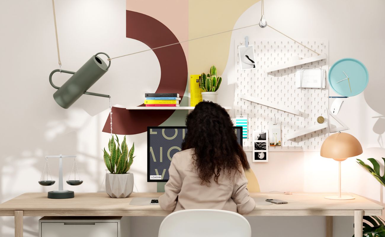 A woman with curly hair sits at a modern desk facing a computer. The workspace features plants, books, a lamp, a wall clock, and creative wall décor with geometric patterns and shelves.