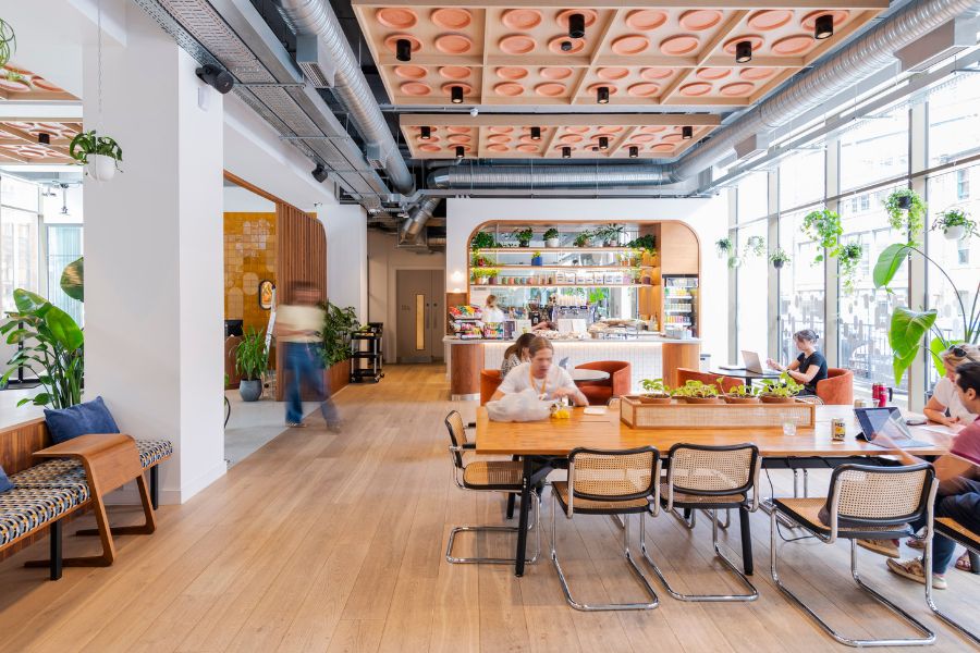 Modern open office space with large windows, wooden floors, and plants. People sit at a long table working and talking, while others move around. A coffee bar with shelves and snacks is in the background. Bright and inviting atmosphere.