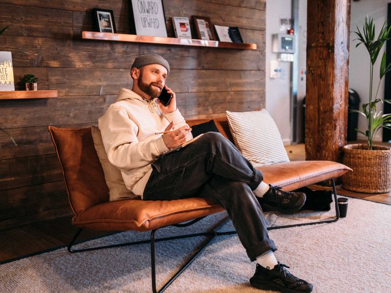 A man with a beard, wearing a beanie and hoodie, sits on a leather couch, talking on his phone and holding a notebook and pen. The modern room has wood-paneled walls, shelves with books, and plants.