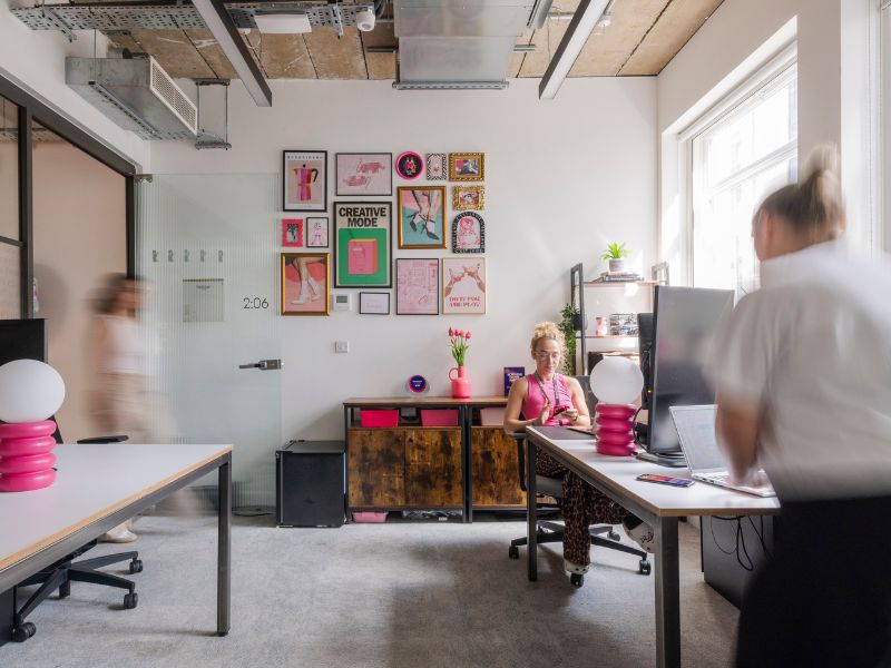 A modern office with colorful decor, showing a woman working at a desk, another person walking past, and a third person in motion. The room has large windows, wall art, and contemporary furniture.