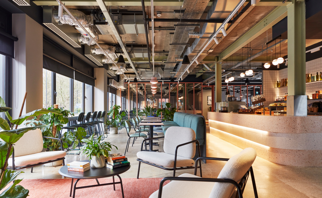 Modern open-plan café with comfortable chairs, round tables, potted plants, large windows, and a counter with warm lighting. Ceiling shows exposed ducts and pipes, creating an industrial yet inviting atmosphere.