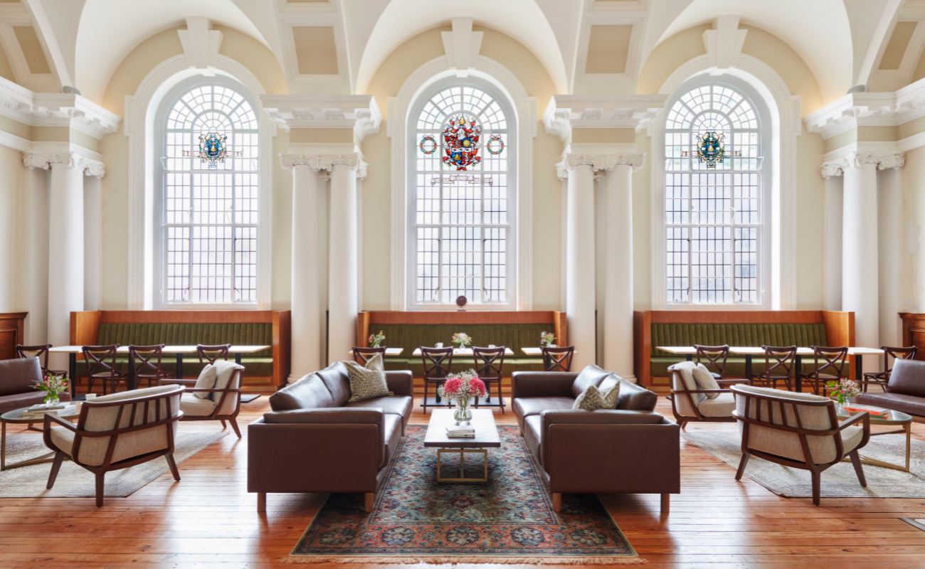 Spacious lounge with high arched windows, elegant columns, stained glass details, brown leather sofas, armchairs, wooden tables, and a patterned rug on polished wood floors. Natural light fills the room.