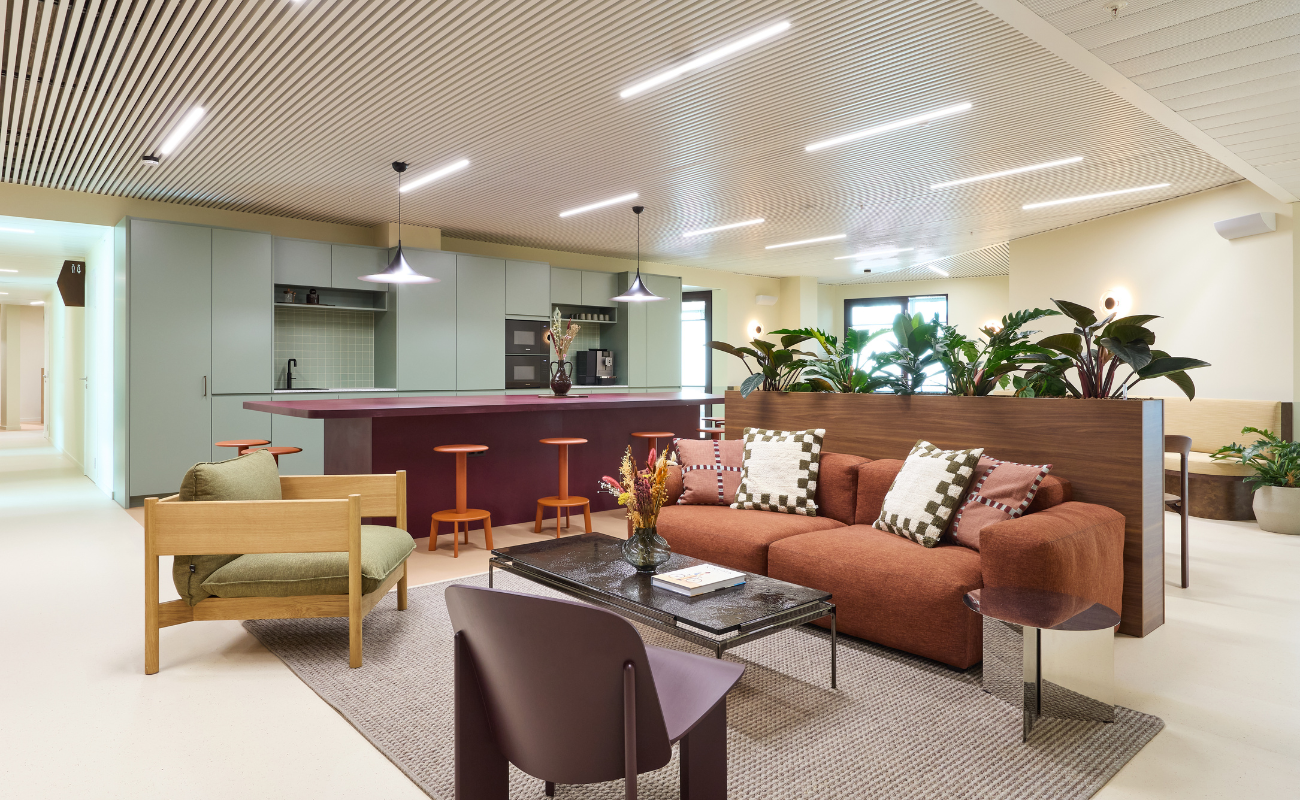 Modern lounge area with a rust-colored sofa, patterned cushions, wooden chairs, a rug, a coffee table, and a kitchen in the background with green cabinets, bar stools, and pendant lights; plants are placed throughout the space.