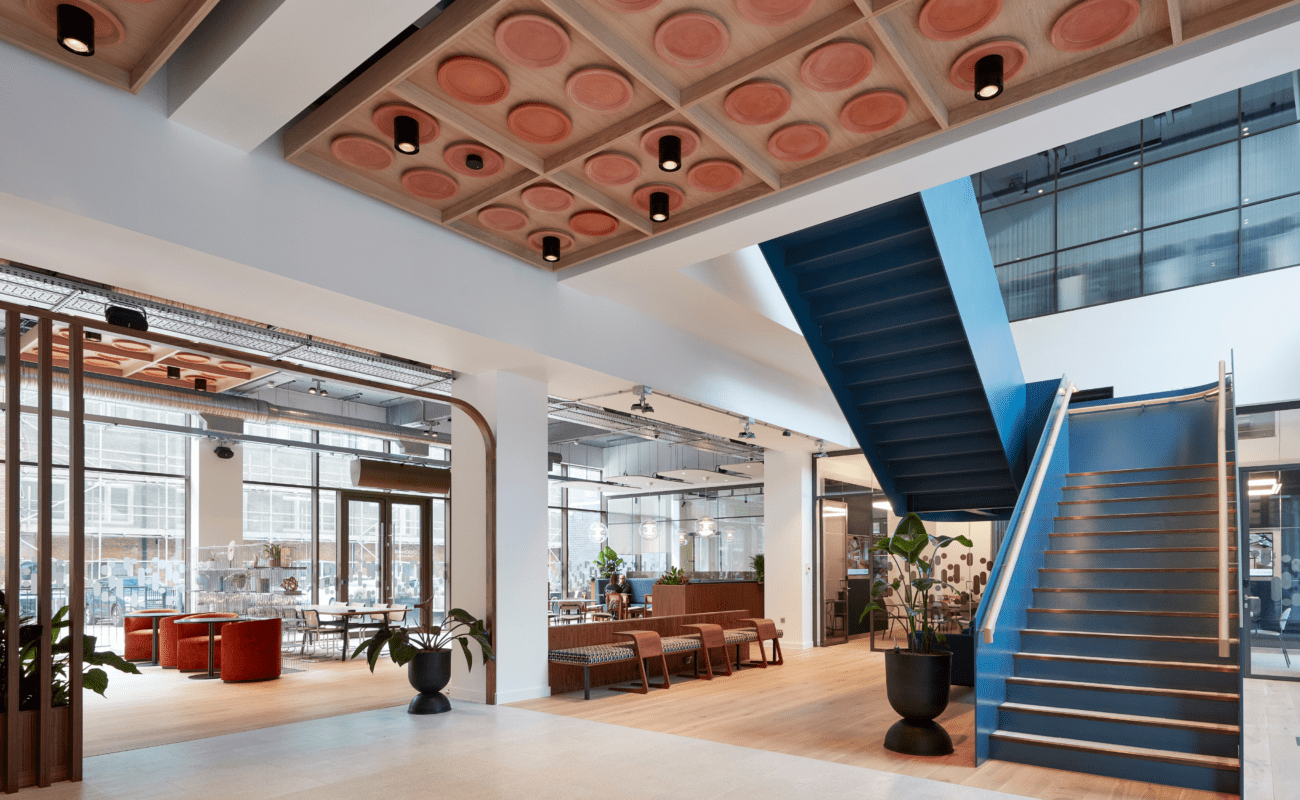 Modern open office space with a blue staircase, wooden ceiling panels, large windows, lounge seating, and potted plants. Natural light fills the area, creating a bright and welcoming atmosphere.