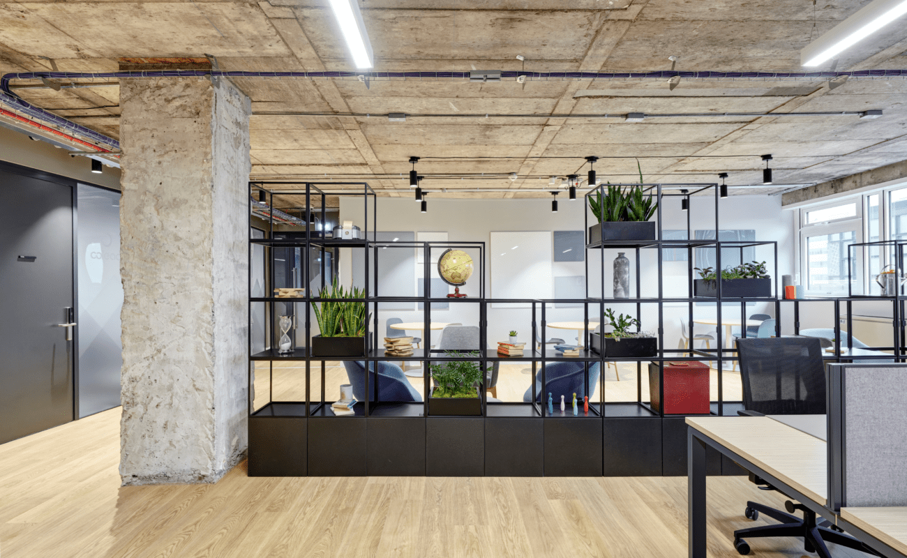 Modern open-plan office space with industrial design elements, featuring an exposed concrete ceiling, partition shelves with plants and decor, wooden floors, and natural light from large windows.