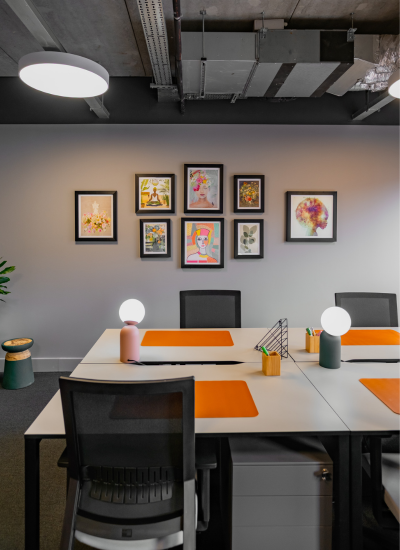 A modern office space with desks, black chairs, orange desk mats, round desk lamps, and colorful artwork in black frames on a gray wall. Exposed ceiling and minimal decor create a contemporary atmosphere.