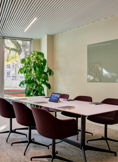 A modern conference room with six maroon chairs around a white table, a large plant by the window, notebooks and a tablet on the table, and a glass board mounted on the wall. Natural light fills the space.