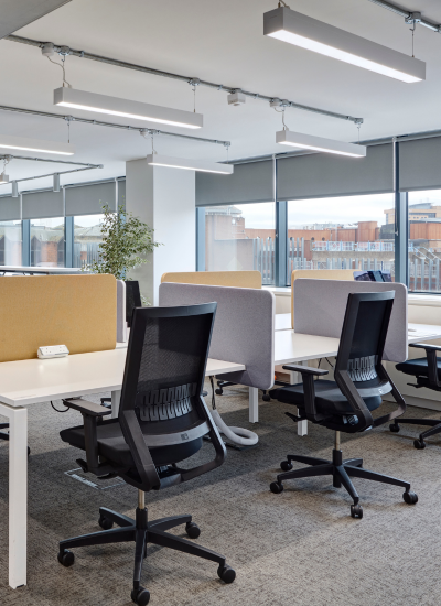 A modern open-plan office with several empty desks and ergonomic chairs, each workstation separated by fabric partitions. Large windows provide natural light and a view of buildings outside.