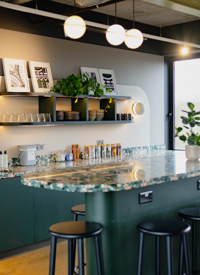 A modern kitchen with a green marble countertop, black stools, open shelves holding dishes and framed art, potted plants, and globe pendant lights hanging from the ceiling.