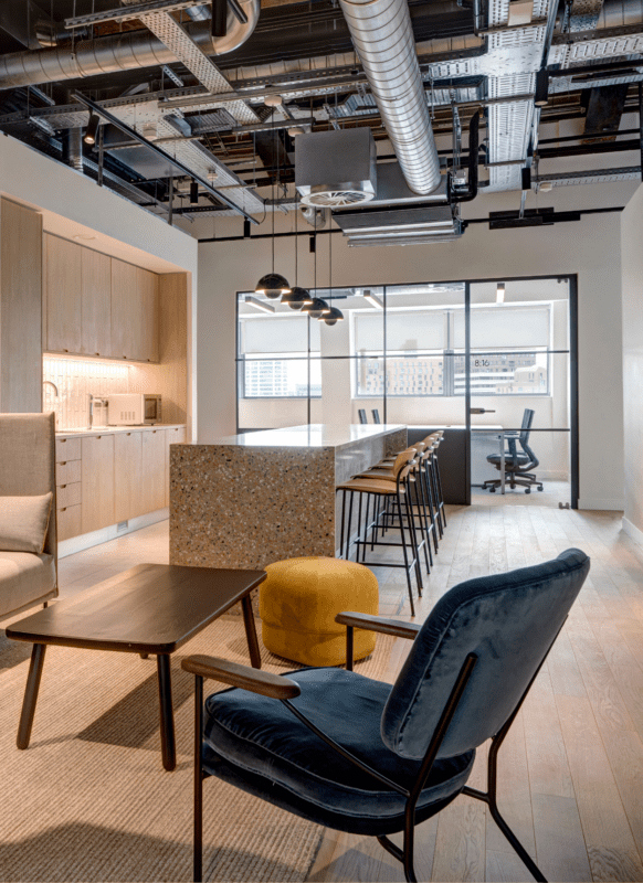 Modern office lounge with a blue velvet chair, yellow ottoman, wooden table, and a kitchen area featuring light wood cabinets, a terrazzo island with barstools, and large windows showing city buildings.