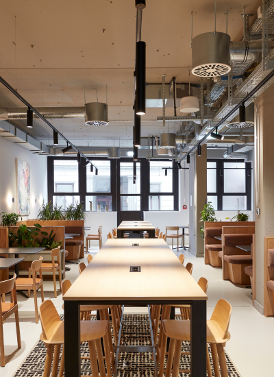 A modern, well-lit office café with wooden tables and chairs, brown booth seating along the walls, potted plants, and large windows letting in natural light. Industrial ceiling fixtures are visible above.