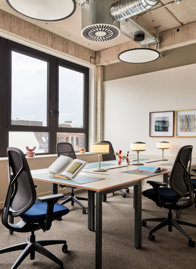 A modern office space with four workstations, each with a black chair, desk lamp, and open book. Large windows let in natural light, and framed art hangs on neutral walls. Exposed ceiling pipes are visible above.
