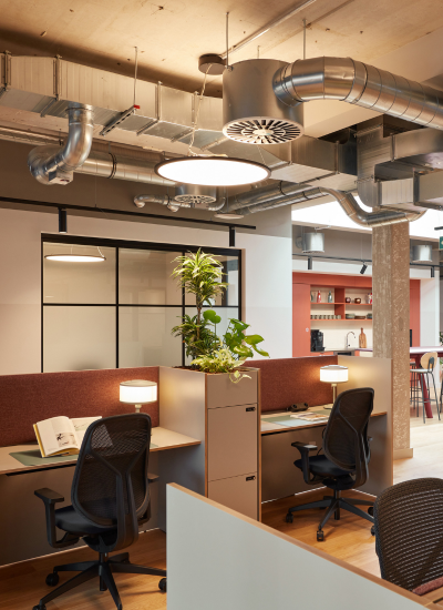 Modern office space with two workstations, ergonomic chairs, desk lamps, an open book, and a plant partition. Exposed ductwork and a kitchenette area are visible in the background.