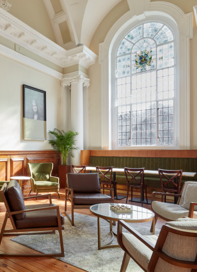 A bright, elegant room with tall windows, wooden floors, modern chairs, a large arched window with stained glass, a framed portrait, potted plant, and comfortable seating arranged around a glass coffee table.
