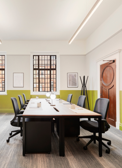 Modern office with a long desk, six black chairs, minimalist lamps, large windows with black frames, framed art on pale green and white walls, and a wooden door. The space is bright and clean.