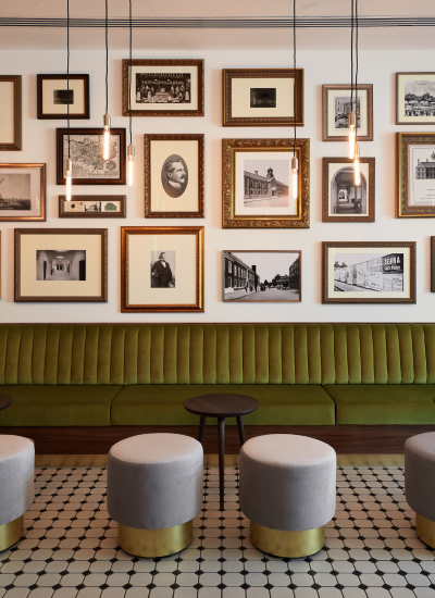 A modern lounge with green upholstered seating, round gray stools with gold bases, small black tables, and a wall decorated with framed vintage photos and artwork under hanging pendant lights. Black and white tiled floor.