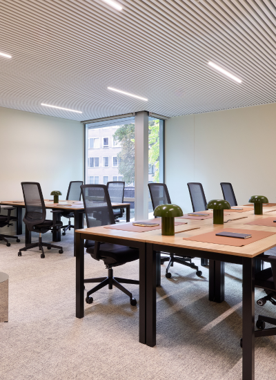 Modern office space with wooden desks arranged in rows, each equipped with a green desk lamp, black mesh chairs, and notebooks. Large window in the background lets in natural light and shows trees and a building outside.