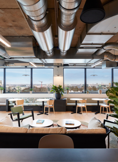 A modern office lounge with large windows, exposed ductwork, light wood furniture, green plants, and a view of the city skyline on a clear day.