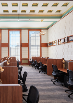 A spacious, well-lit room with rows of wooden study cubicles, black chairs, table lamps, large tall windows, and framed pictures on the wall, featuring high ceilings with decorative molding and a chandelier.