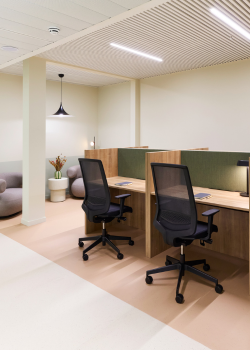 Modern office space with two workstations separated by wooden partitions, each with a mesh office chair, desk lamp, and notepad. In the background, there is a seating area with armchairs, a side table, and a hanging light.
