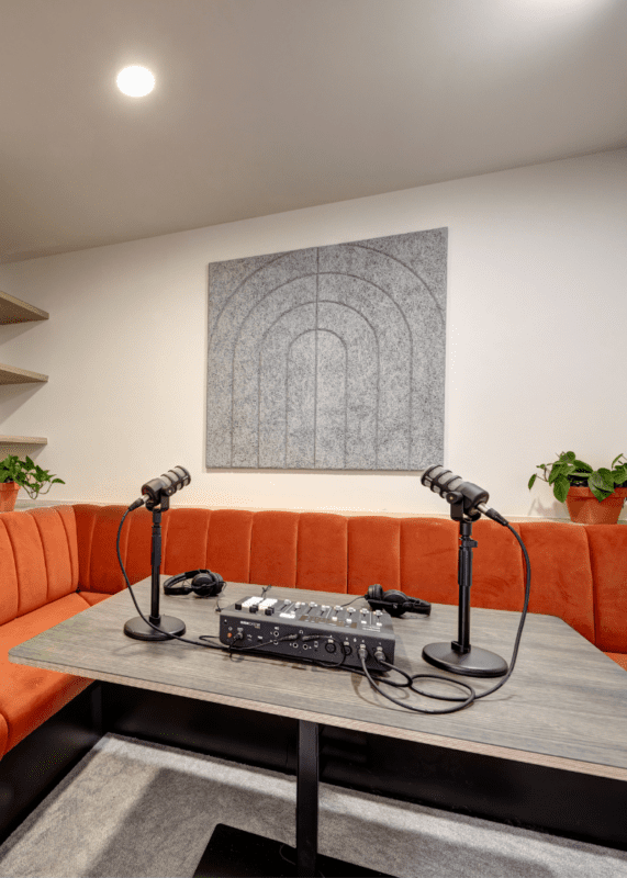 A modern podcast recording setup with two microphones, headphones, and audio equipment on a table, surrounded by orange upholstered seating and potted plants, beneath abstract wall art.