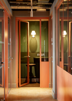 A modern phone booth with orange-framed glass walls stands open in a hallway, revealing a black chair, a small desk, and a wall-mounted lamp inside, with soft lighting and green acoustic panels on the walls.