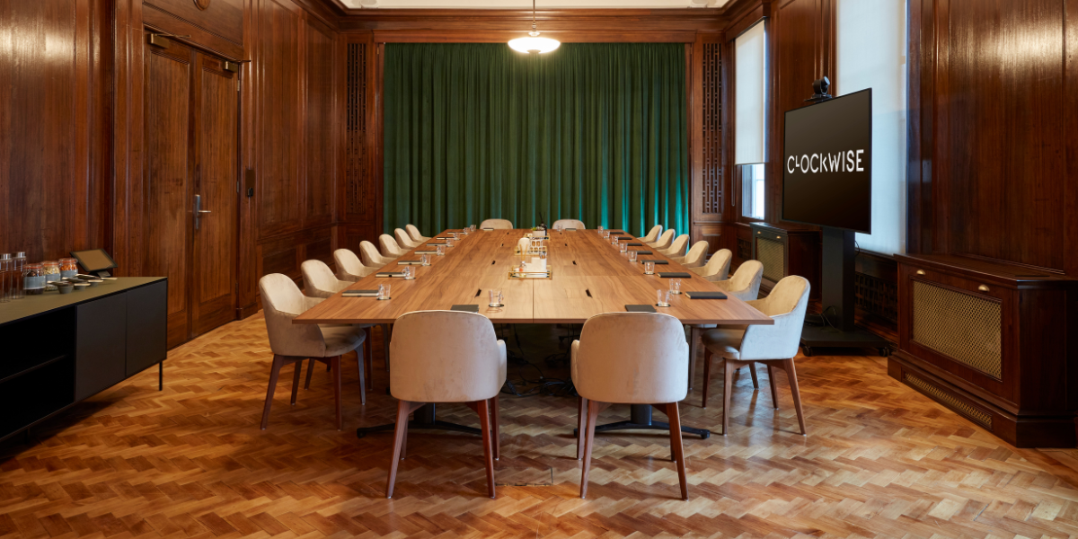 A boardroom with a long wooden table surrounded by white chairs, set with notepads and glasses. The room has wood-paneled walls, parquet flooring, a green curtain, and a large screen displaying 