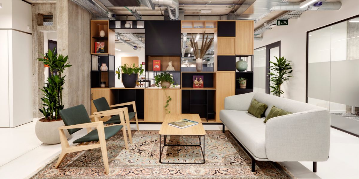 A modern office lounge with a light gray sofa, two wooden chairs with green cushions, a wooden coffee table, a patterned rug, potted plants, and a decorative shelving unit with books and vases.