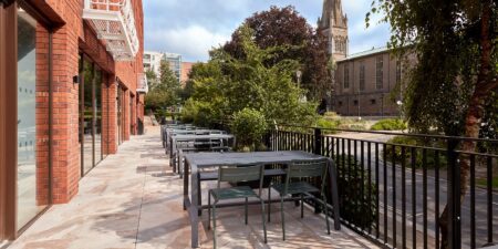 Outdoor patio with several empty tables and chairs next to a brick building, overlooking a garden with trees and a church in the background. The area is peaceful and well-maintained.