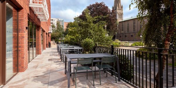 Outdoor patio with several empty tables and chairs next to a brick building, overlooking a garden with trees and a church in the background. The area is peaceful and well-maintained.