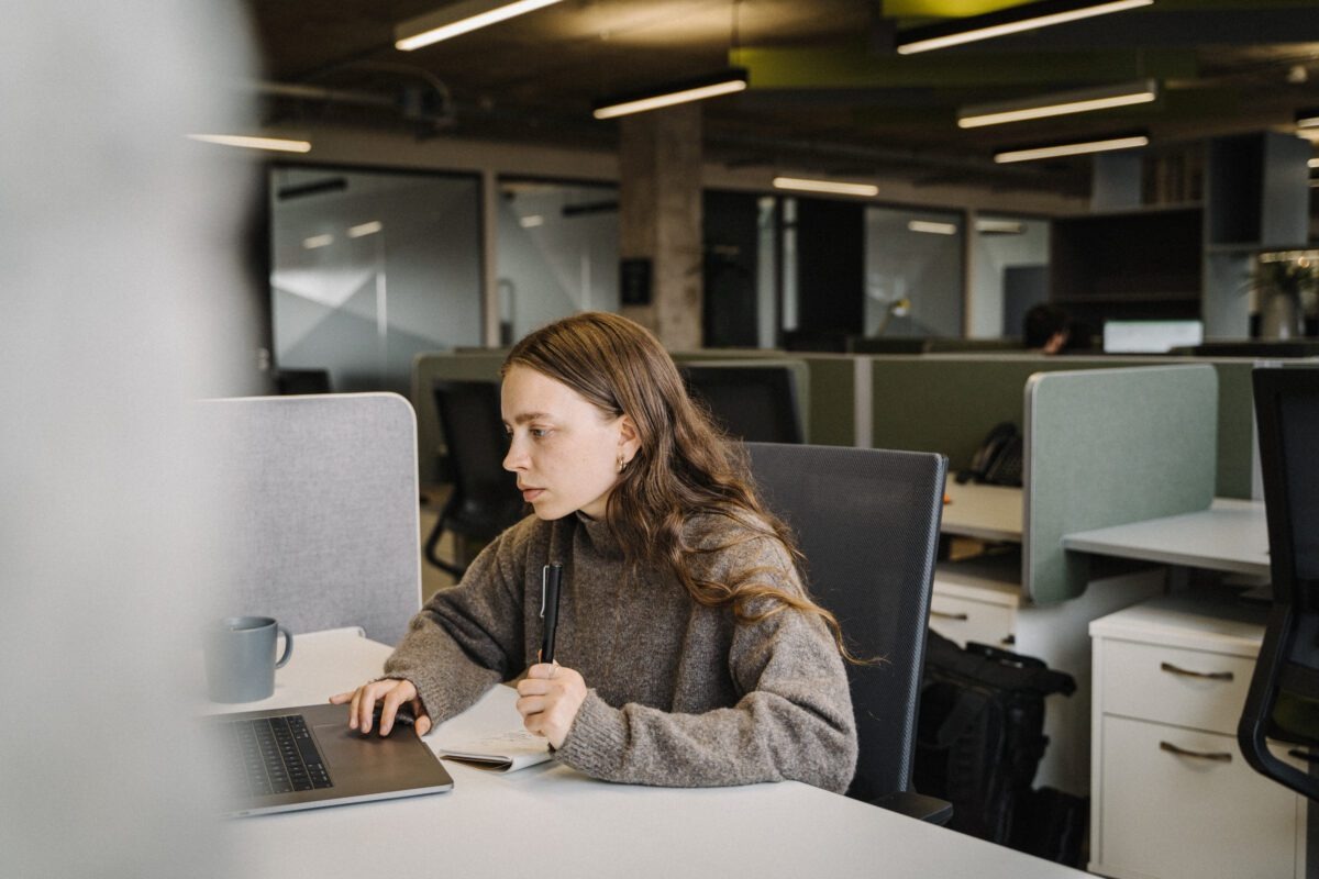 A woman with long brown hair sits at a desk in a modern office, looking intently at her laptop while holding a pen. She wears a grey sweater and has a mug nearby. The office has partitioned workspaces.