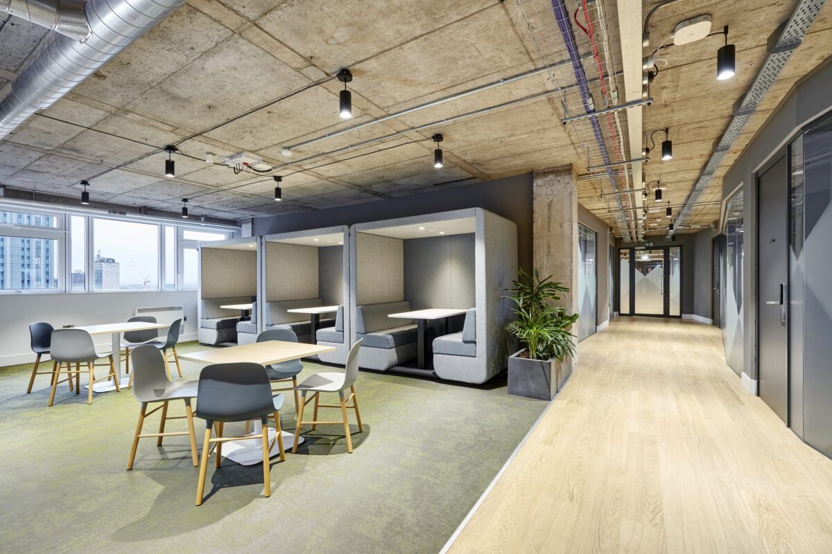 Modern office space with booth seating, tables, chairs, and large windows. The ceiling is exposed concrete with visible pipes. There are potted plants along the wall and hallway access on the right.