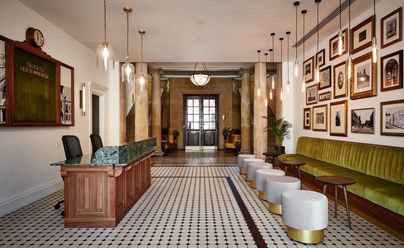 Elegant hotel lobby with a marble check-in desk, green velvet bench, round stools, pendant lights, vintage photos on the wall, and tiled floor leading to double doors framed by columns.